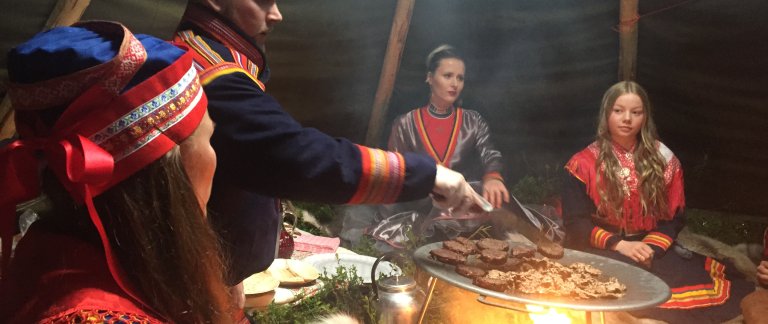 4 people taking part in a Sami Experience sitting around a big plate on a bonfire inside a tent.